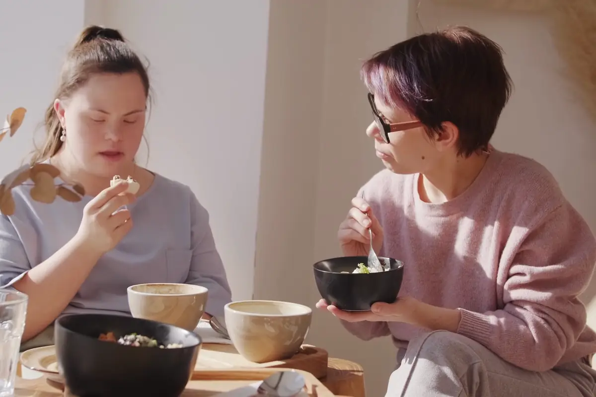 A support worker and a young woman with Down syndrome sharing a meal at a sunny table